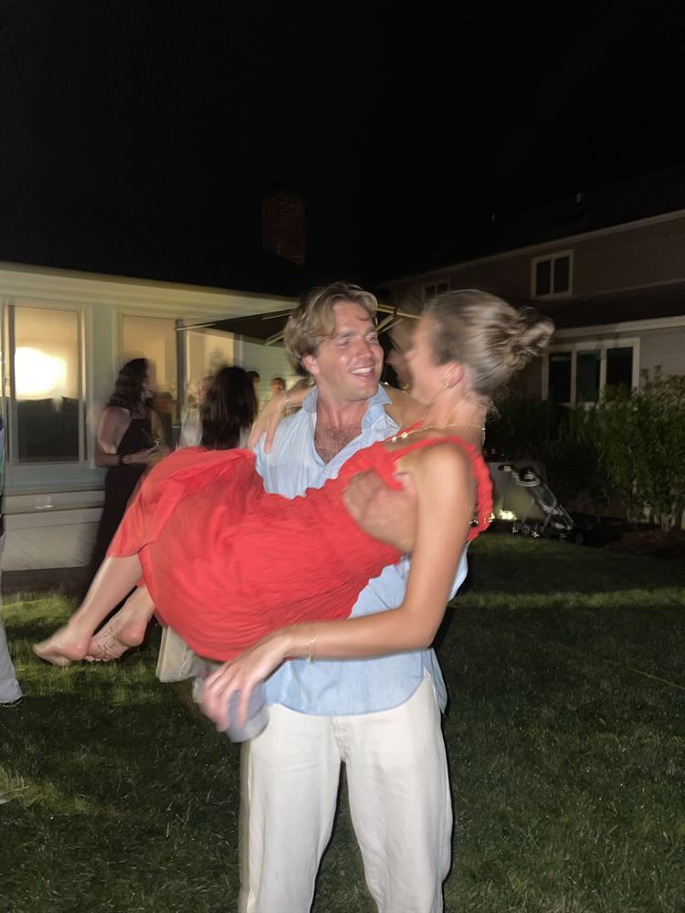 Blake carrying Cami across a lawn at a summer party